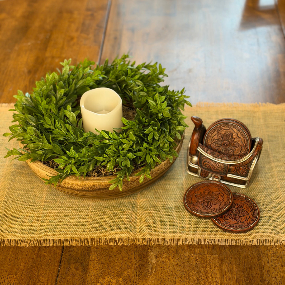 A rustic home decor arrangement on a wooden table featuring a green boxwood wreath with a white pillar candle, next to a small carved wooden chair and two matching carved coasters on a woven runner.