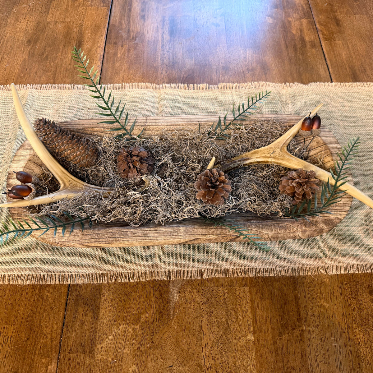 A rustic table centerpiece featuring two antlers, pinecones, acorns, and green pine needles arranged on a natural wood board over gray moss and burlap, placed on a wooden dining table.