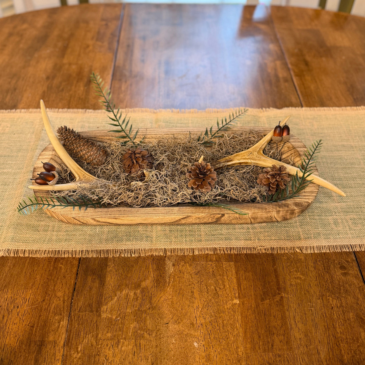 A rustic Christmas table centerpiece in a wooden tray, filled with natural moss, pinecones, evergreen branches, and antlers, displayed on a burlap runner over a wooden dining table.