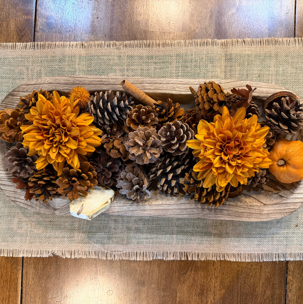 A rustic wooden serving tray filled with autumn decorations, including two large orange dahlia flowers, pinecones, a small orange pumpkin, and a cinnamon stick, placed on a beige burlap runner over a wooden floor.