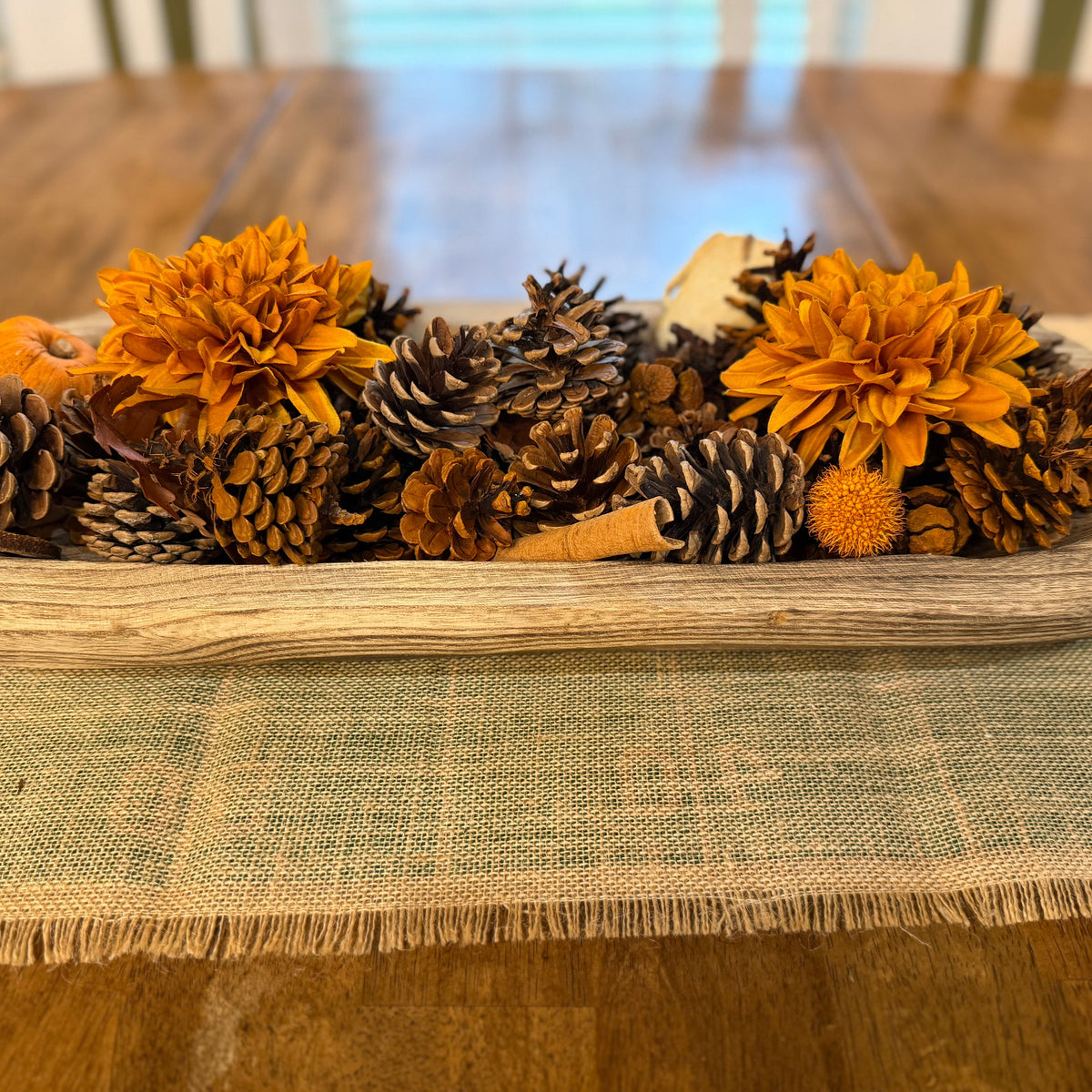 A rustic wooden tray filled with dried orange flowers, pinecones, and a small orange pumpkin, displayed on a woven table runner over a wooden dining table.
