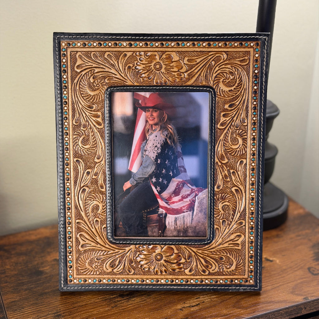 A rectangular leather picture frame with ornate hand-tooled floral detailing and a black border, displayed on a wooden table against a neutral wall.