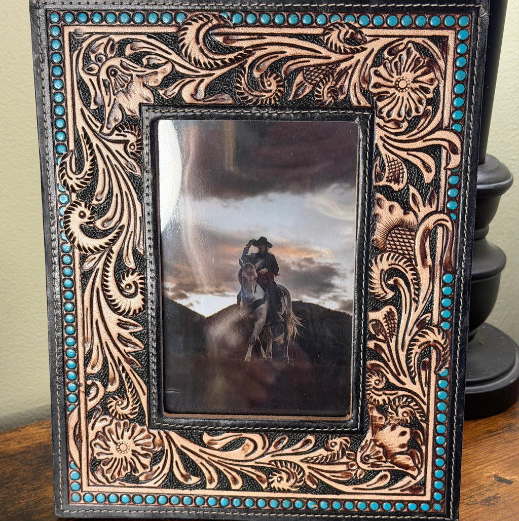 A hand-tooled leather picture frame with intricate floral and scroll designs, turquoise bead accents, and a central photo of a cowboy on horseback, displayed on a wooden surface.