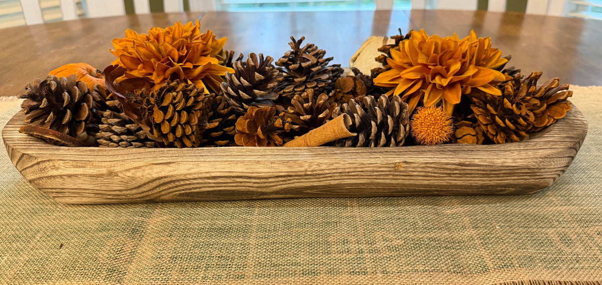 A rustic wooden tray filled with dried orange flowers, pinecones, and small orange berries, displayed on a woven table runner over a wooden dining table with chairs in the background.