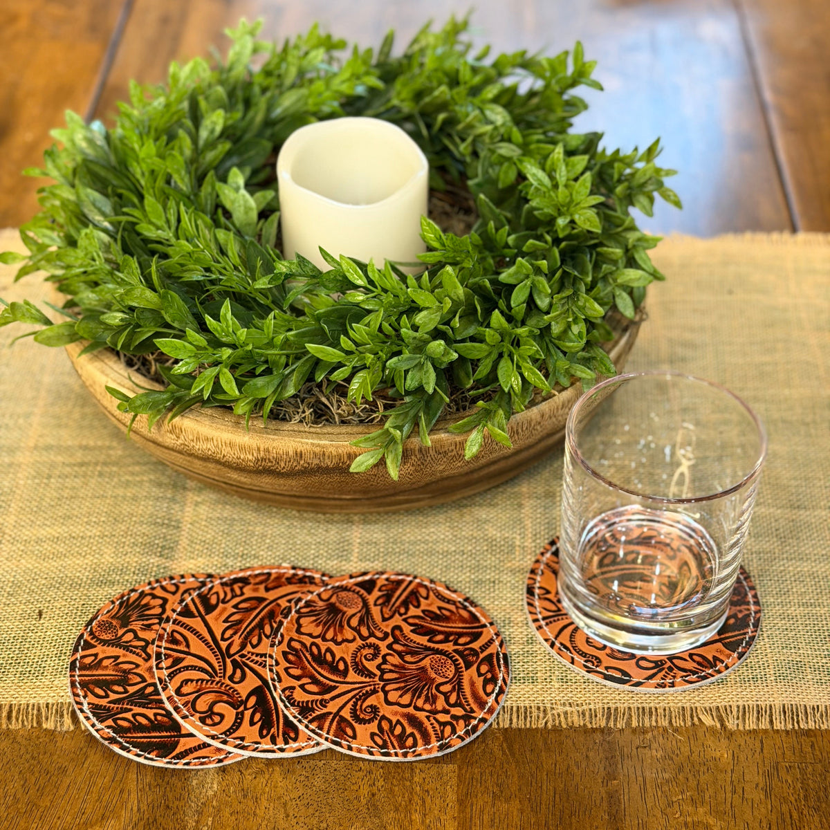 A set of round coasters with an orange and black paisley pattern, shown on a wooden table with a glass and a green wreath centerpiece in the background.