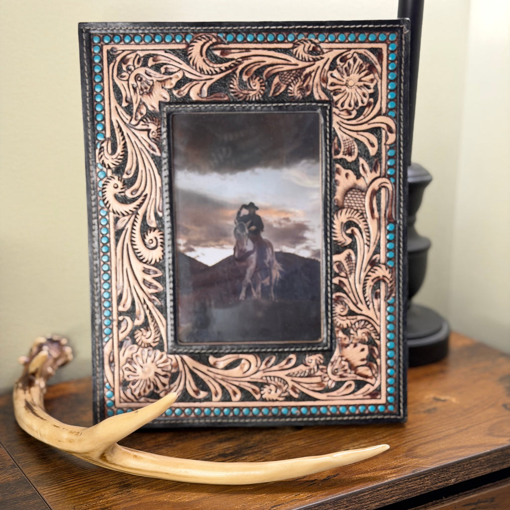 A hand-tooled leather picture frame with intricate floral carvings and turquoise accents, displayed on a rustic wooden cabinet next to a horn antler.
