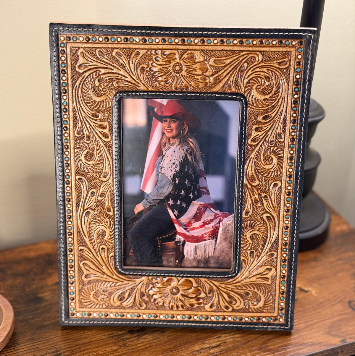 A rectangular hand-tooled leather photo frame with ornate floral carving and small colorful bead accents, displayed on a wooden surface. The center is filled with a sample photo of a woman in a cowboy hat.