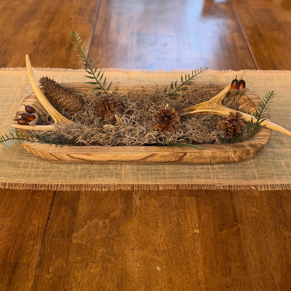 A rustic pinecone and antler centerpiece arranged in a shallow wooden tray with evergreen sprigs and moss, placed on a woven runner over a wooden dining table.