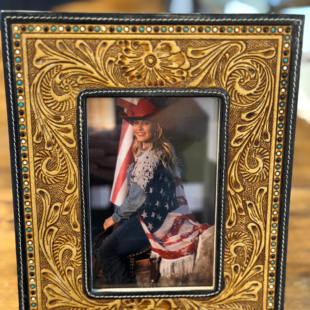 An ornate rectangular picture frame with embossed floral detailing and small rhinestone accents, shown standing on a wooden surface. The center insert contains a portrait photo of a woman in western attire holding an American flag.