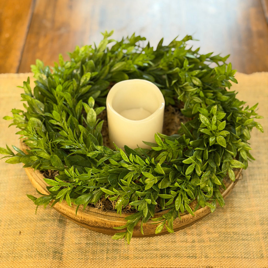 A green leafy wreath with a white pillar candle holder in the center, displayed on a burlap-covered surface with a wooden table in the background.