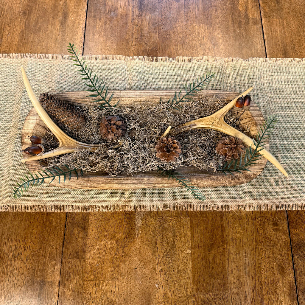 A rustic holiday table centerpiece featuring natural antlers, pinecones, evergreen branches, and dried grass on a wooden board, placed on a burlap runner over a wooden dining table.