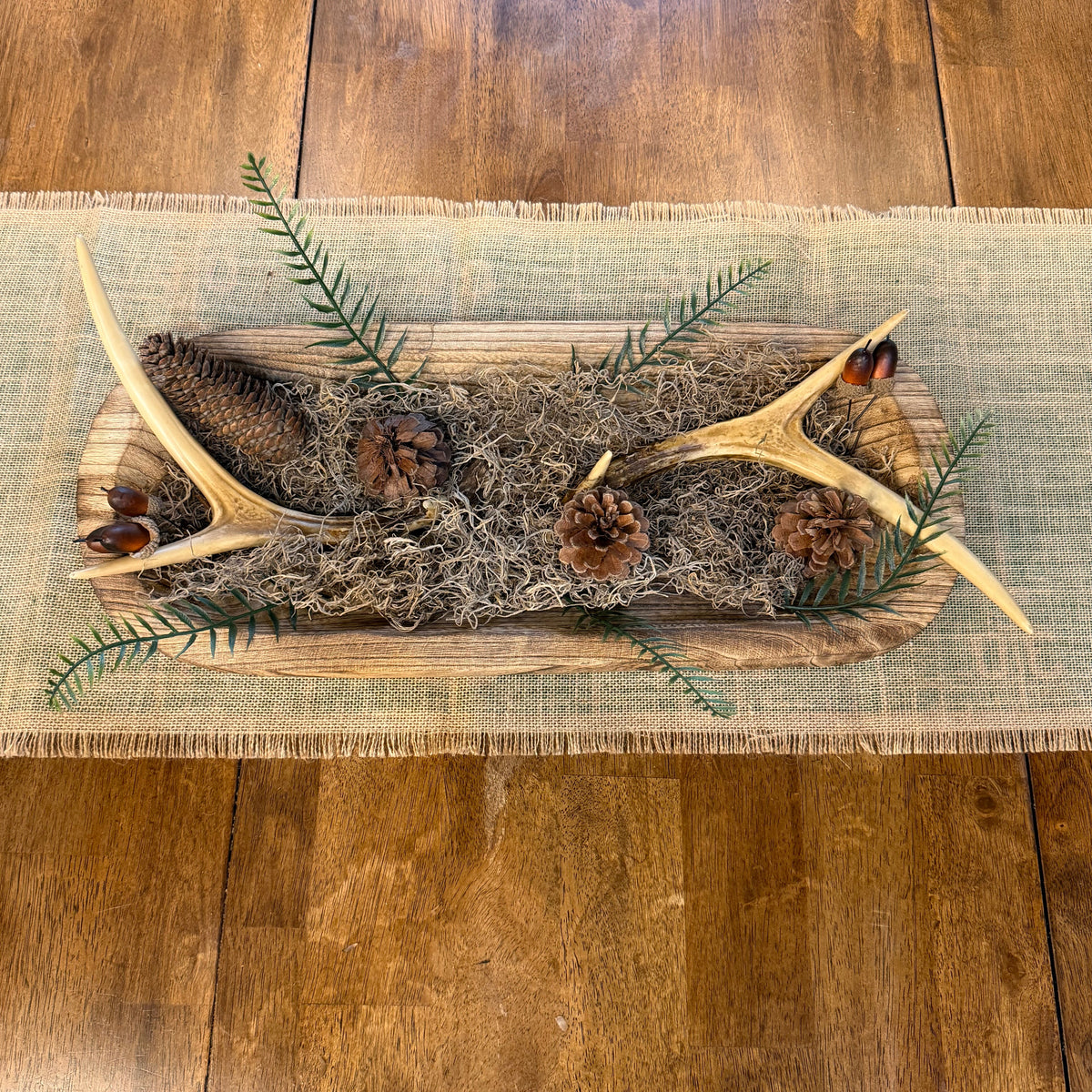 A rustic holiday table centerpiece featuring natural antlers, pinecones, evergreen branches, and dried grass on a wooden board, placed on a burlap runner over a wooden dining table.