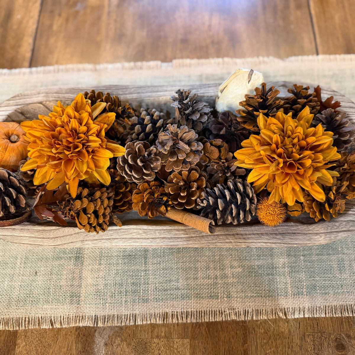 A rustic fall-themed centerpiece in a long oval wooden bowl, filled with orange dahlia flowers, pinecones, a small orange pumpkin, a cinnamon stick, and a white decorative stone, set on a woven placemat over a wooden table.