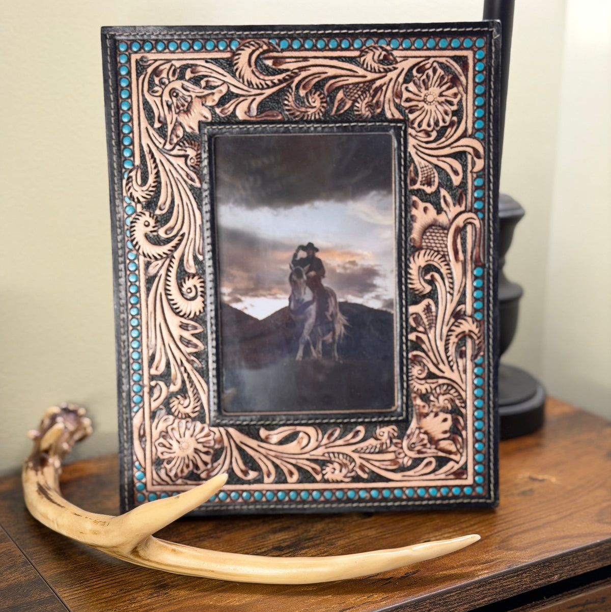 An ornate tooled leather picture frame with turquoise accents, displaying a western-style portrait of a cowboy on horseback, styled on a rustic wooden table next to a horn antler.