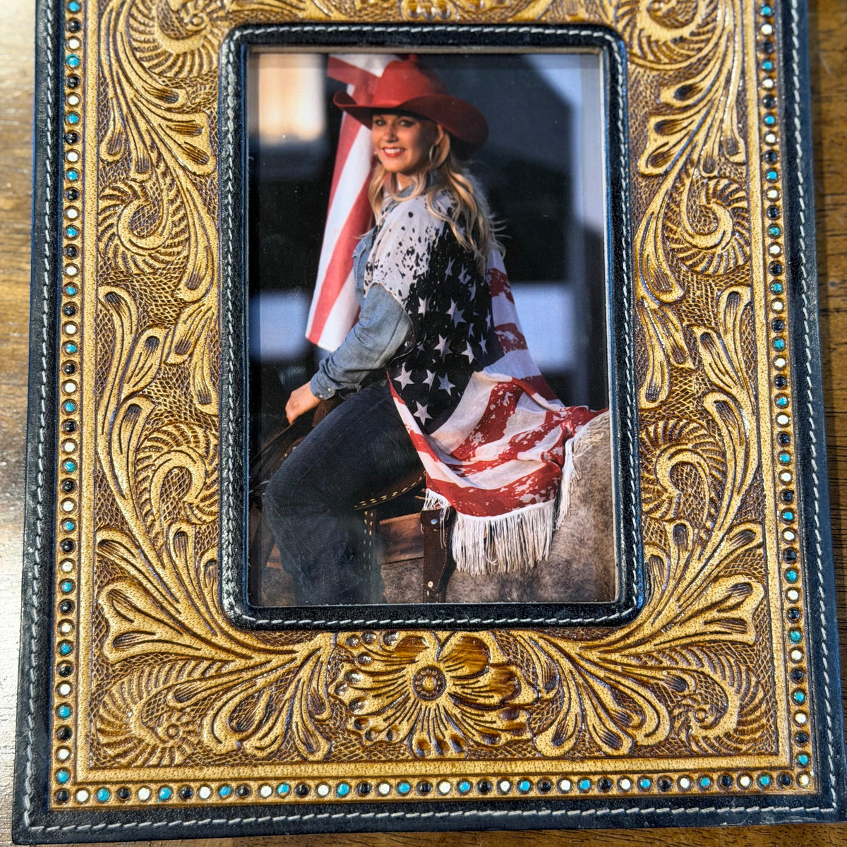 An ornate, hand-tooled leather picture frame with intricate floral carving and small turquoise rhinestone accents, displayed on a wooden surface. The center is filled with a sample photo of a woman in western wear.
