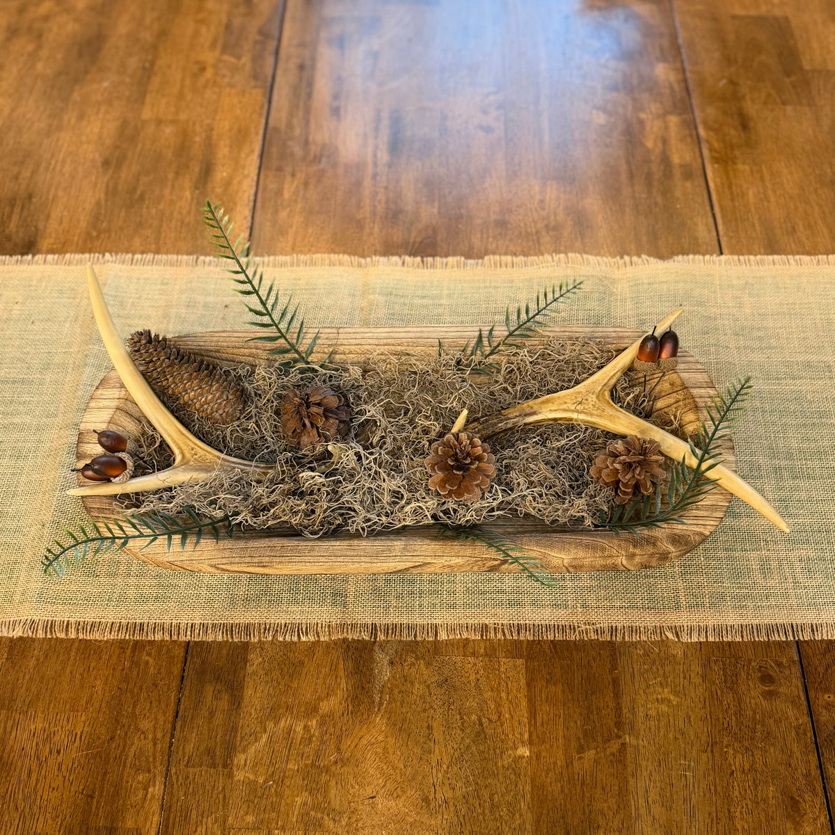 A rustic fall-themed table runner centerpiece on a wooden dining table, featuring natural antlers, pinecones, evergreen branches, and dried moss inside a shallow wooden tray over a beige woven runner.