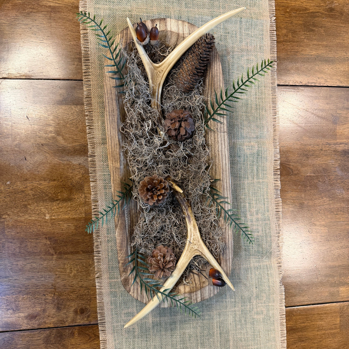 A rustic table runner centerpiece featuring two antlers, pinecones, and green pine branches arranged on a wooden board with gray moss, placed on a light blue burlap runner over a dark wood floor.