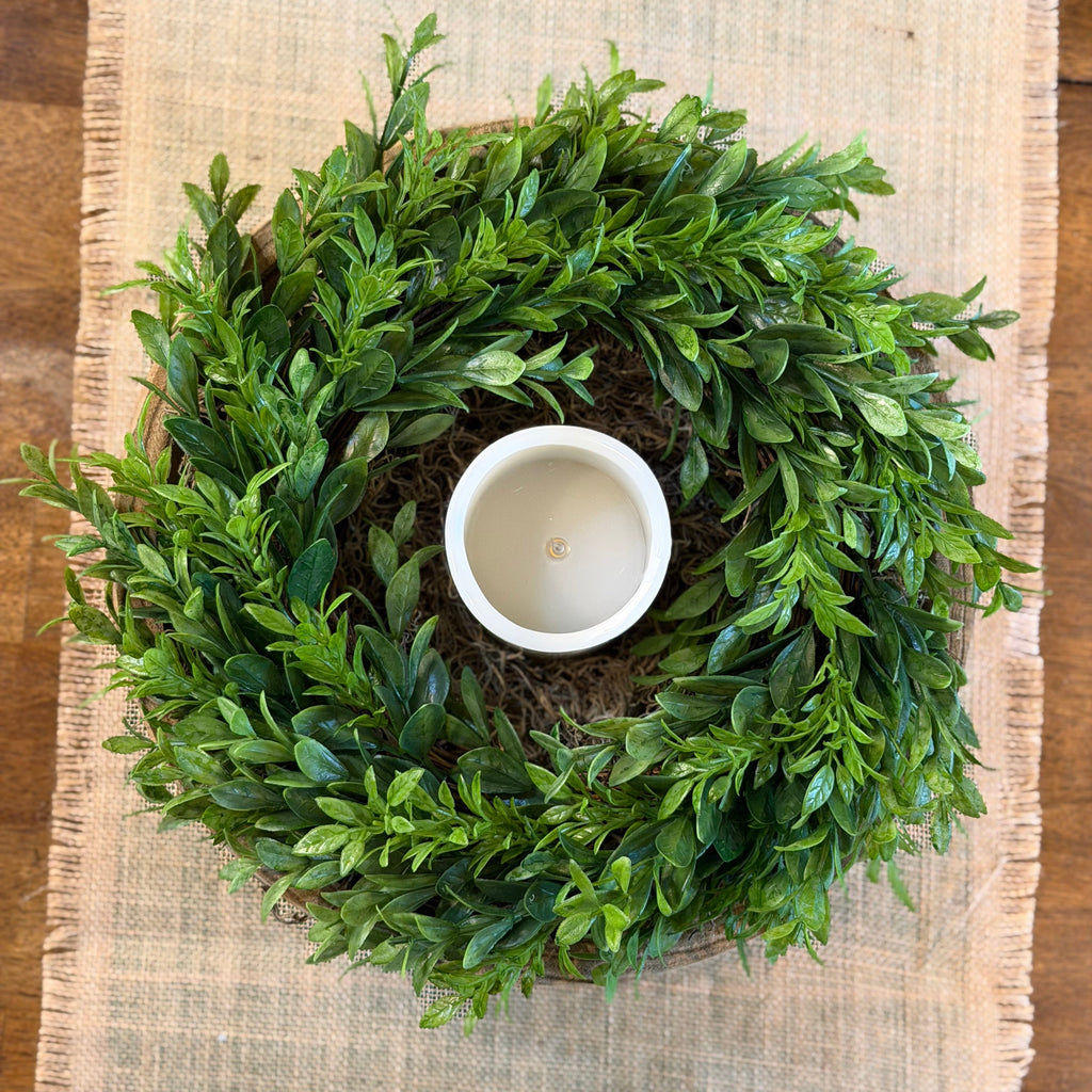 An artificial green boxwood wreath with a white ceramic candle holder in the center, displayed on a beige burlap runner over a wooden surface.