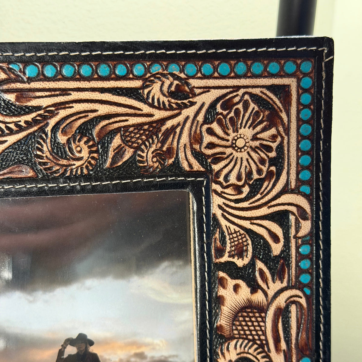 Close-up of a hand-tooled leather picture frame with floral and scroll patterns, turquoise blue bead accents, and a photo insert of a cowboy on horseback.