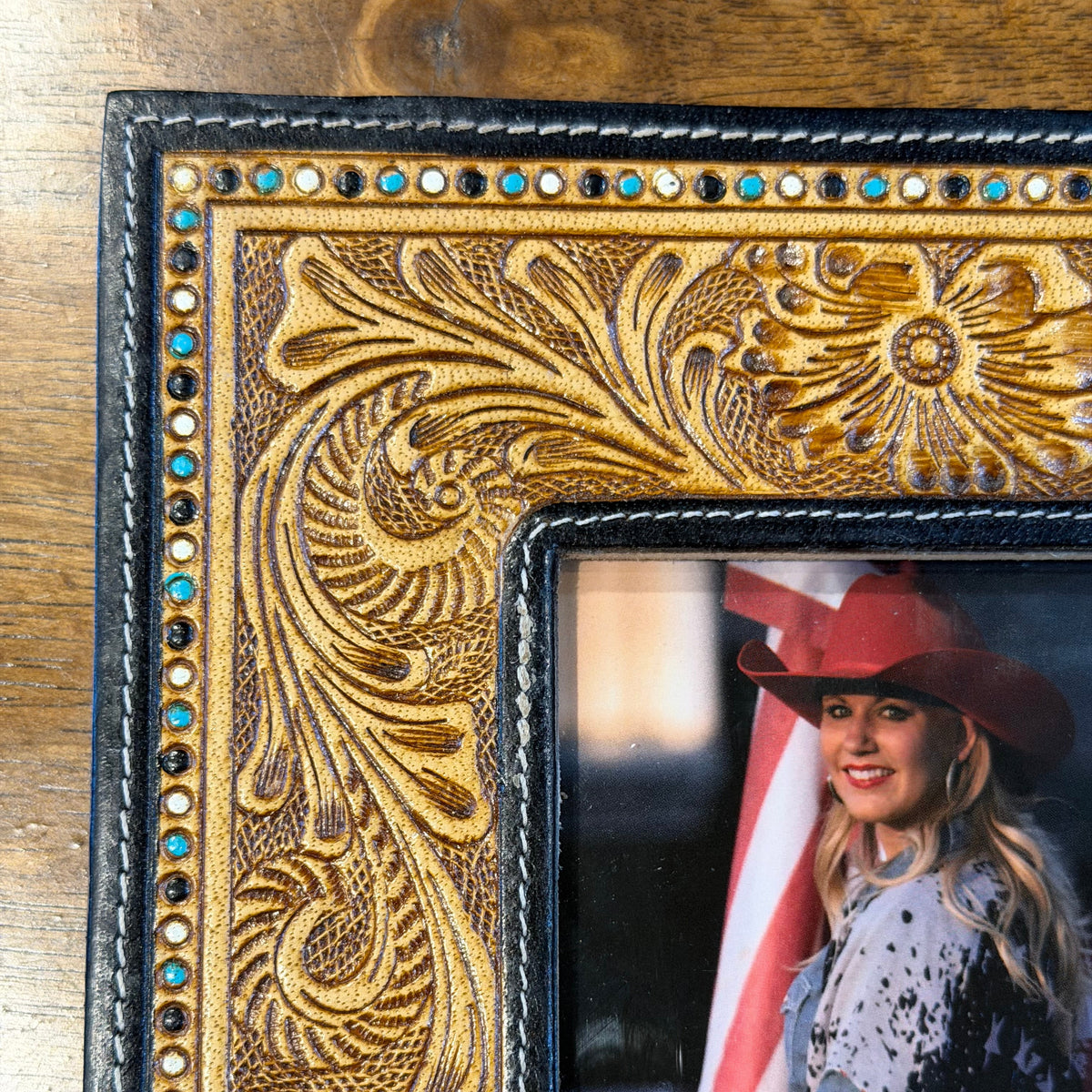 Close-up of a hand-tooled leather picture frame with ornate floral carving and small turquoise and gold rhinestone accents, shown on a wooden surface with a photo insert of a woman in a cowboy hat.
