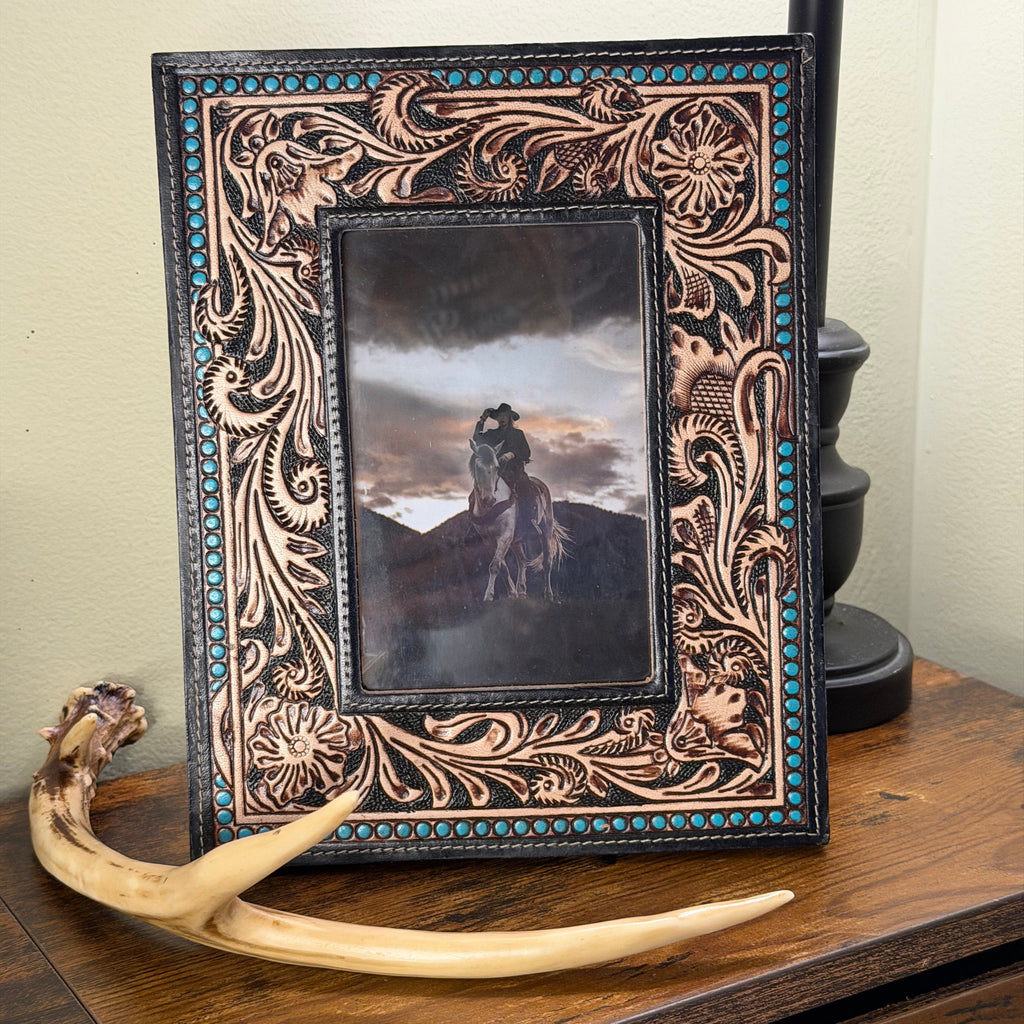 A rectangular hand-tooled leather picture frame with intricate floral carvings and turquoise bead accents, displayed on a wooden table next to a horn antler prop.