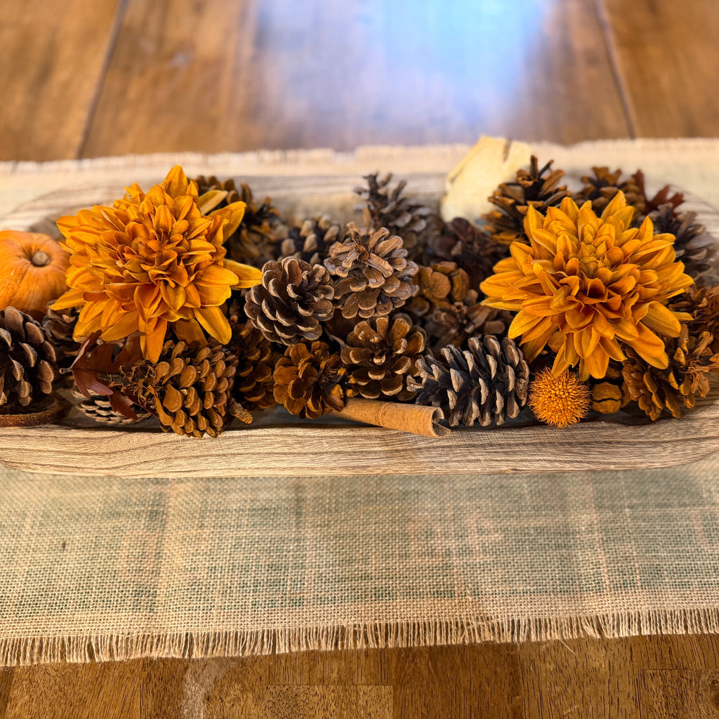 An autumn-themed centerpiece arrangement of orange dahlia flowers, pinecones, a small orange pumpkin, and a cinnamon stick in a rustic wooden oval tray, set on a beige burlap runner over a wooden table.