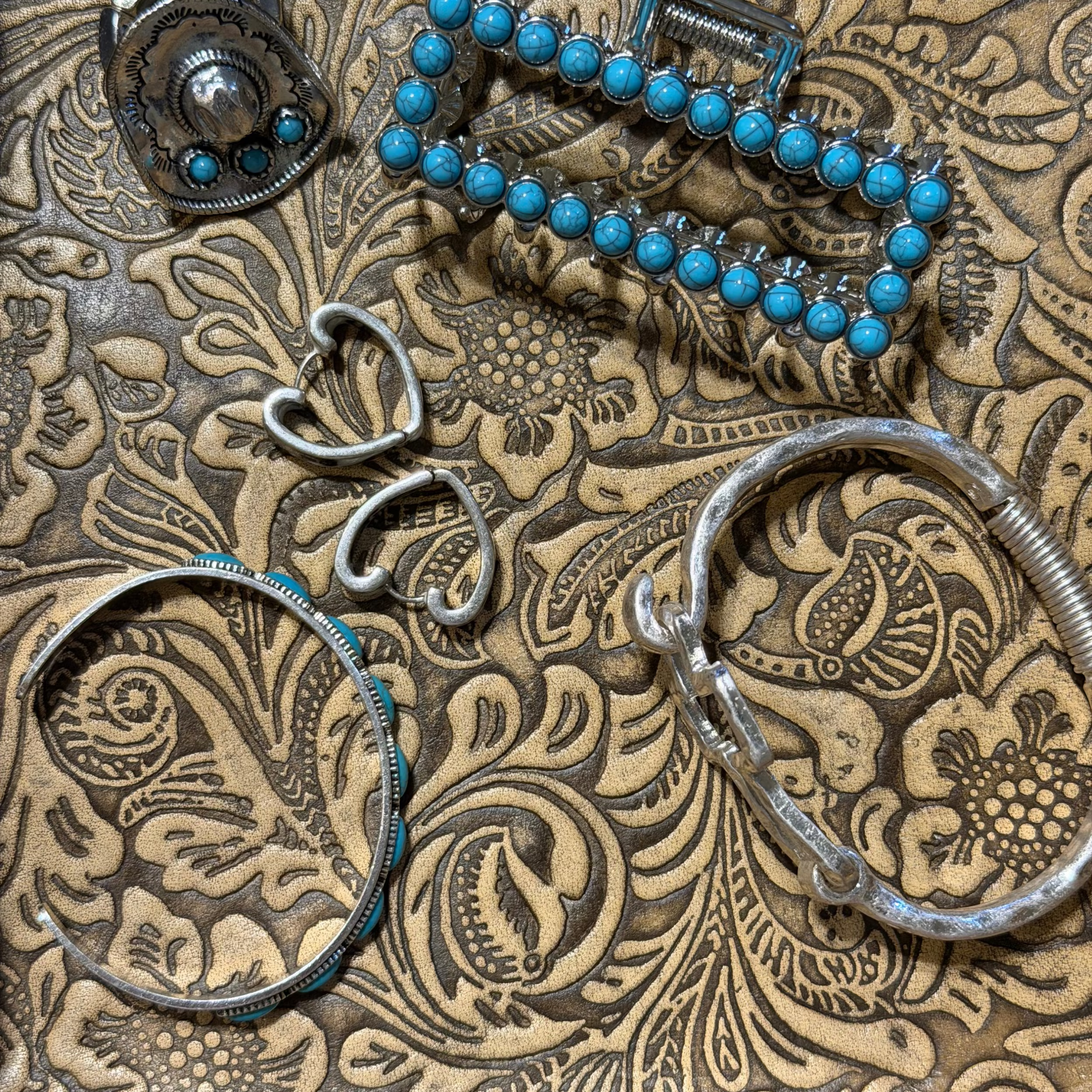 A collection of handmade silver jewelry, including a beaded bracelet, hoop earrings, a ring, and two large cuff-style bracelets, arranged on a heavily embossed brown paisley leather surface.