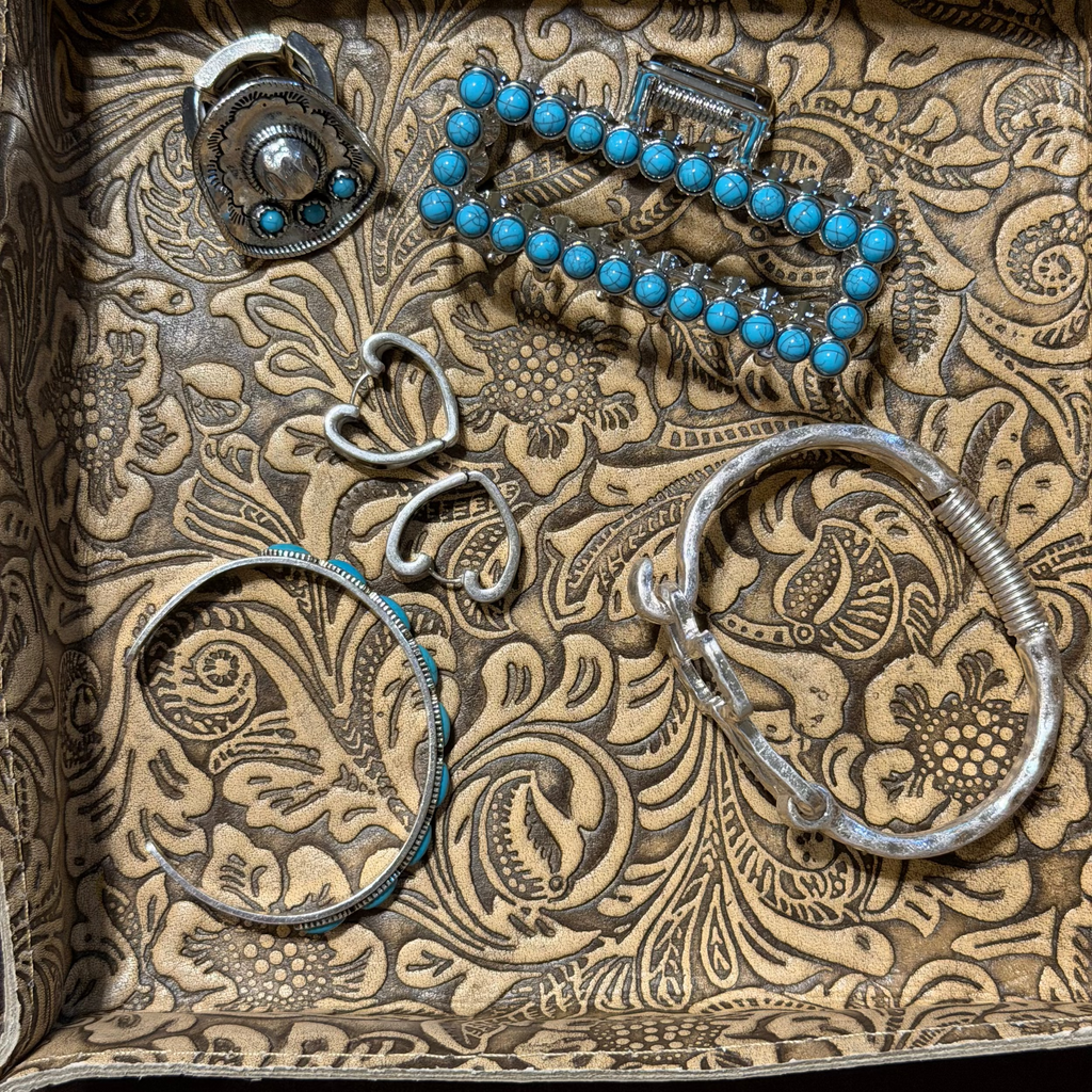 A collection of handmade silver and turquoise jewelry pieces, including bracelets, a ring, and a small heart-shaped charm, arranged inside a decorative brown patterned box.