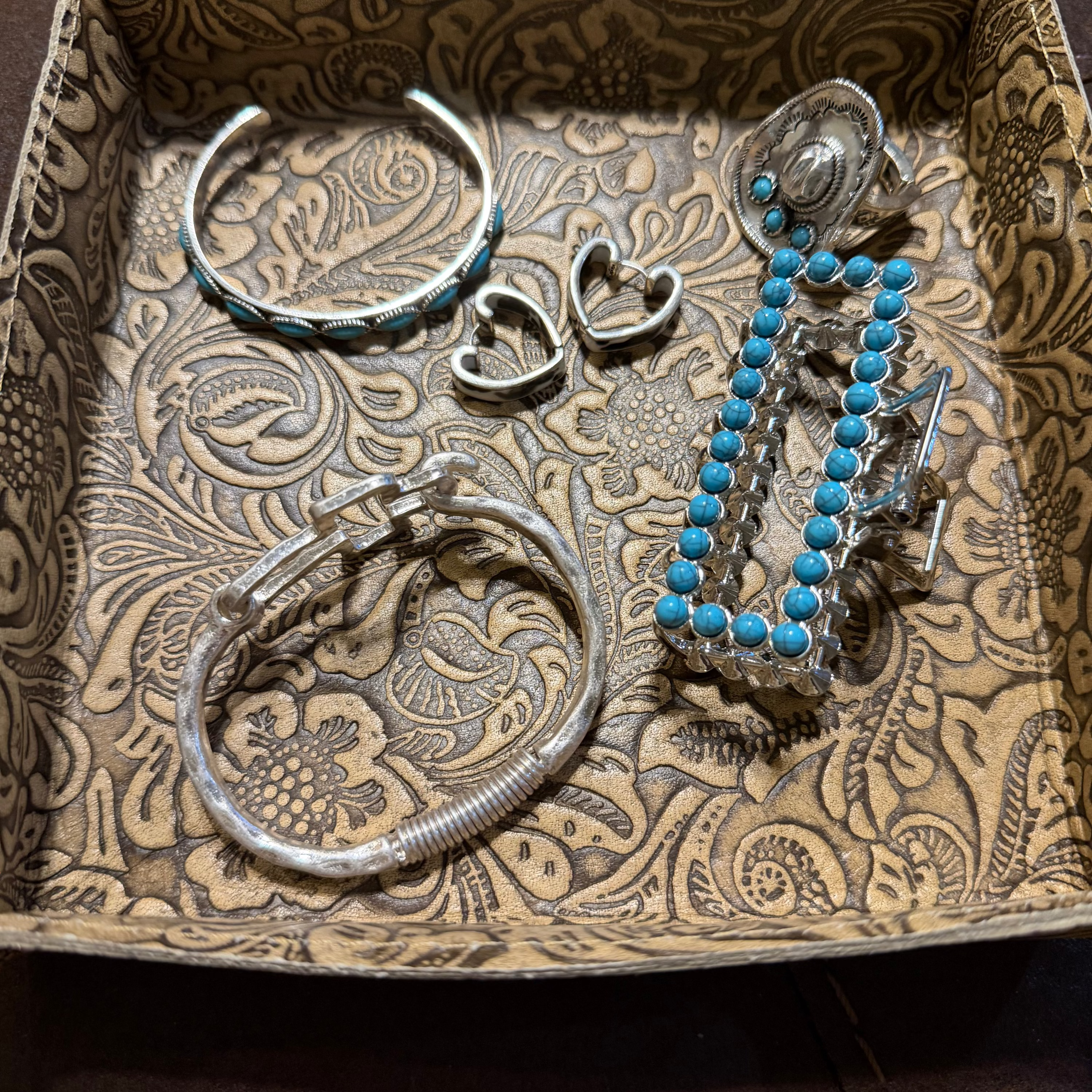 A collection of silver-toned jewelry, including a cuff bracelet, a spring ring, a heart-shaped ring, and a beaded necklace with turquoise blue beads, arranged inside a patterned brown and beige fabric jewelry box.