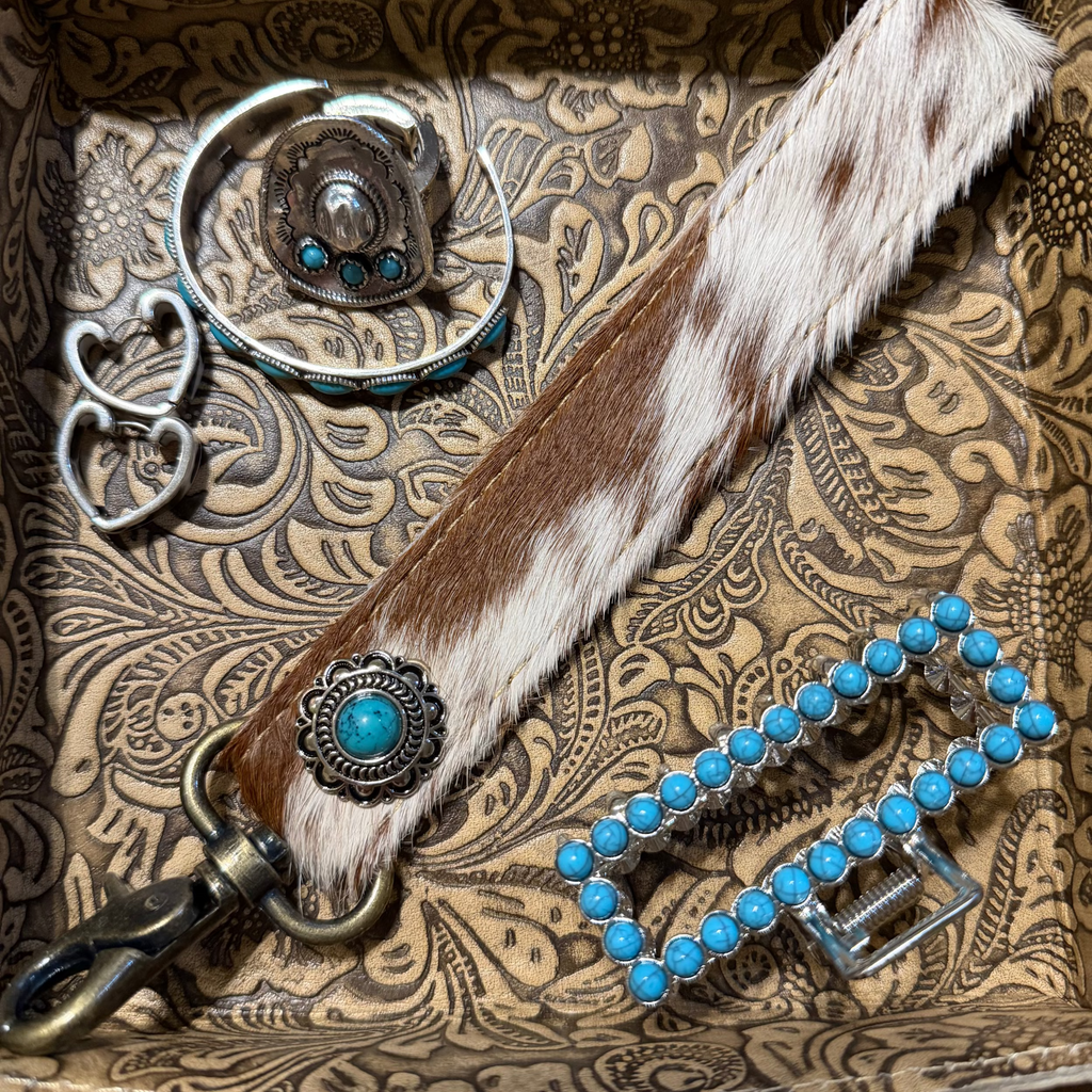 A western-style leather strap with brown and white cowhide pattern, turquoise concho hardware, and a metal clasp, displayed inside a patterned brown tray alongside matching turquoise jewelry.