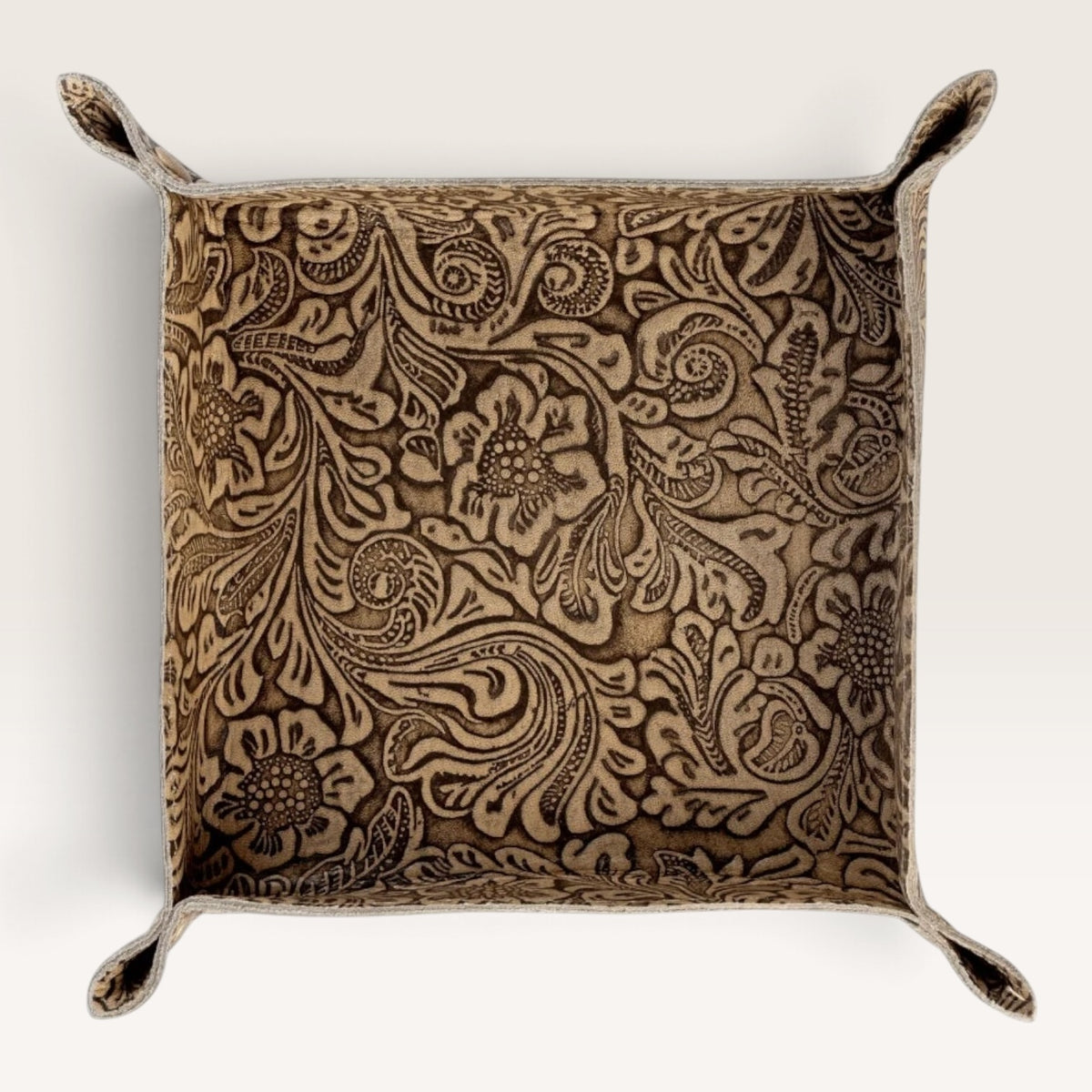 A square decorative pillow with a raised floral and scroll pattern in warm brown and beige tones, featuring stitched loop handles at the corners, shown against a plain off-white background.