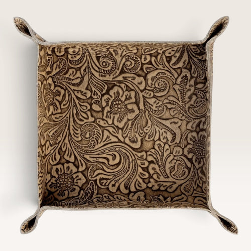 A square decorative pillow with a raised floral and scroll pattern in warm brown and beige tones, featuring stitched loop handles at the corners, shown against a plain off-white background.
