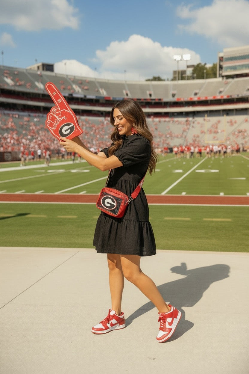 A woman stands on a stadium field holding a red foam finger with a Georgia 'G' logo. She is wearing a black dress, red and white sneakers, and a red crossbody bag with a matching logo patch.