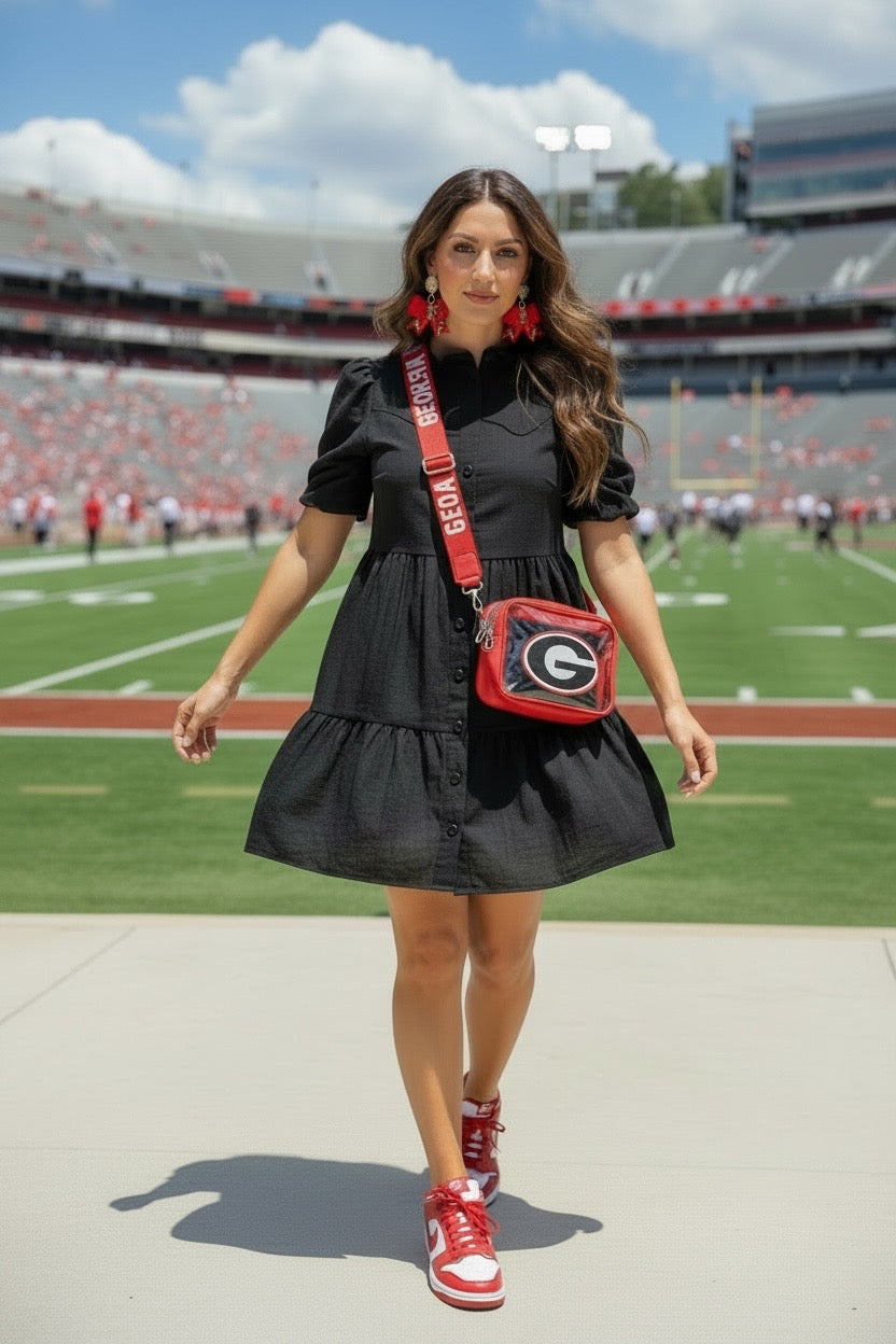 A woman walks on a football field wearing a black tiered mini dress, a red and black Georgia Bulldogs crossbody bag, and red and white sneakers.